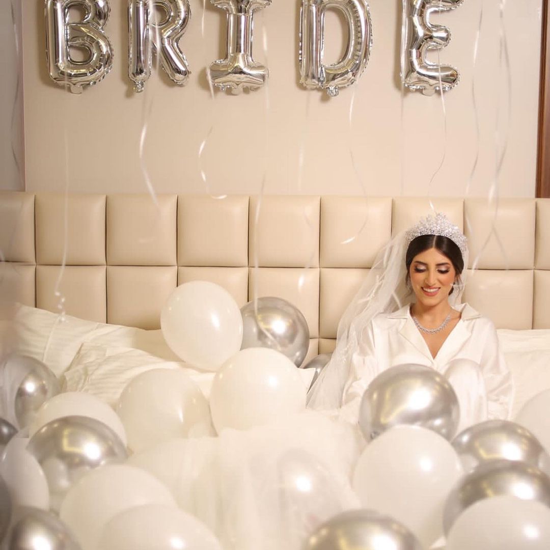 A bride sits on a bed surrounded by white balloons, wearing a white satin pyjama set from Popjammies, radiating elegance and anticipation for her special day.