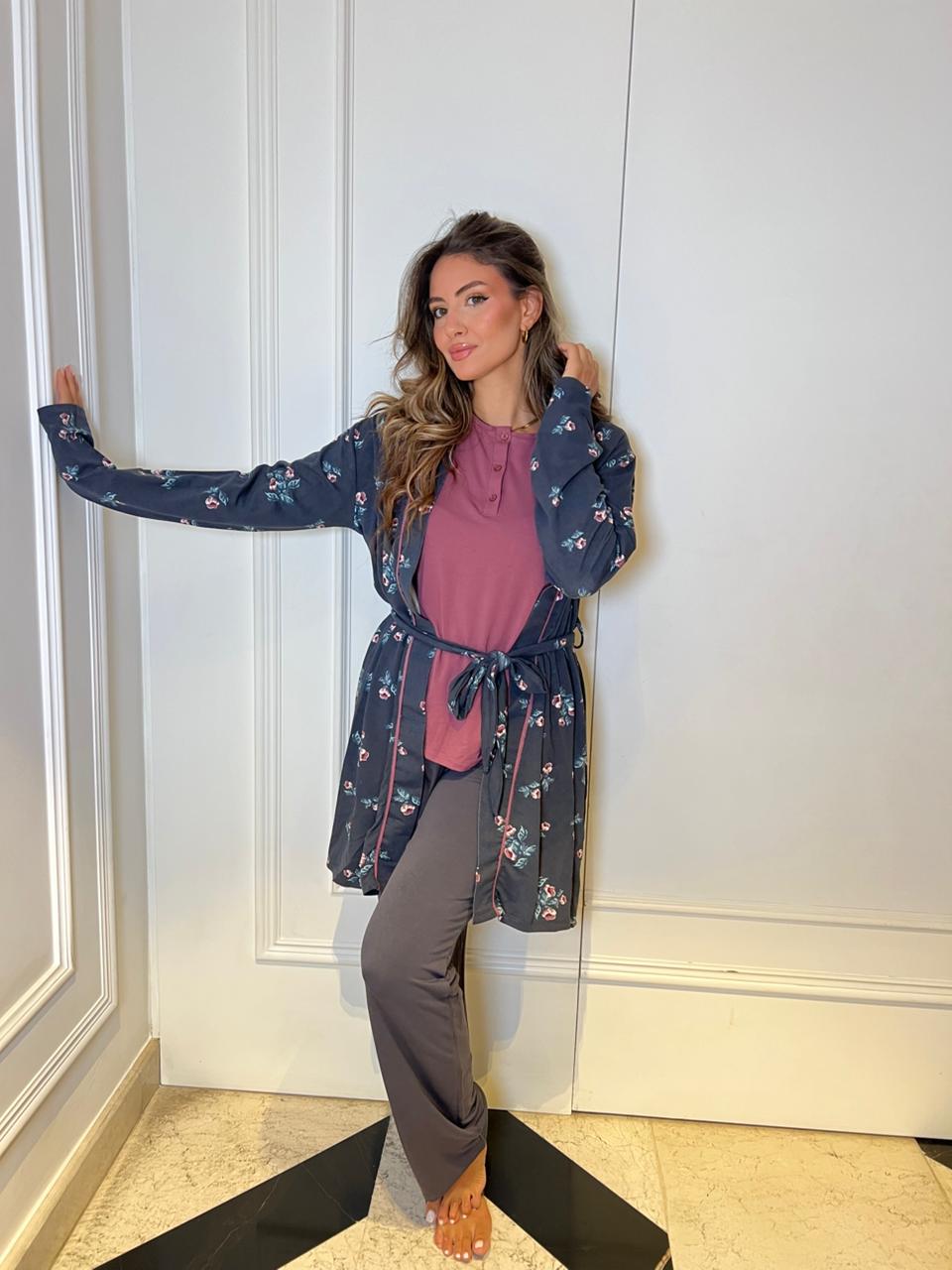Woman wearing a blue floral dress over a pink top and gray pants, standing against a white paneled wall.
