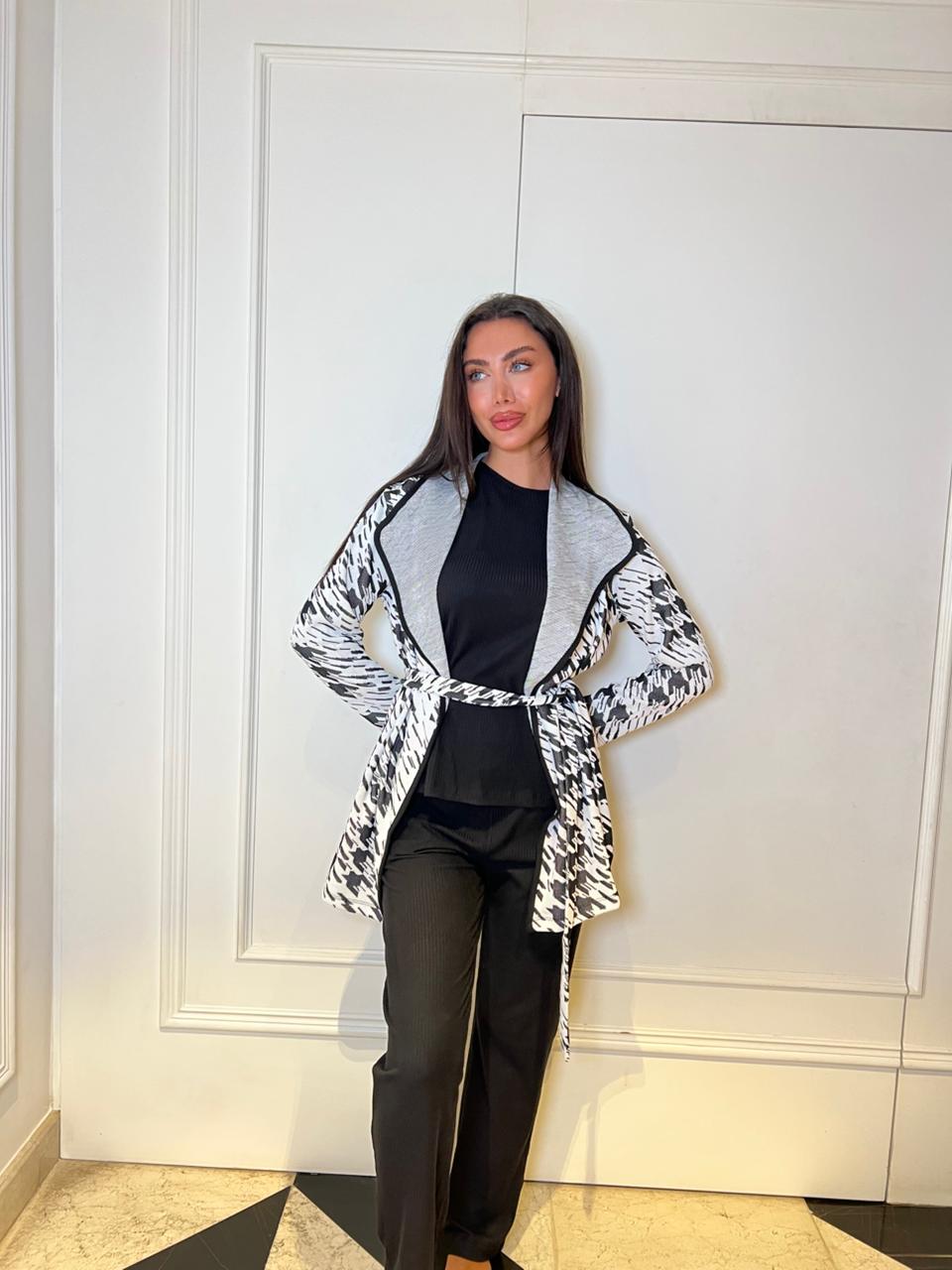 Woman wearing a patterned coat over a black top and pants against a white wall.