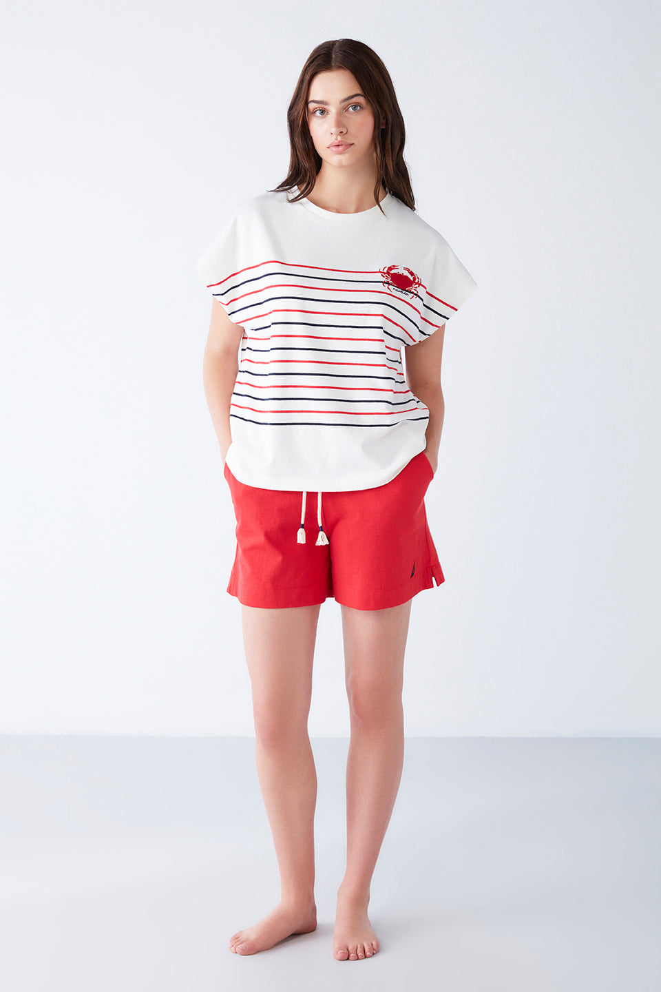 Woman wearing a Popjammies pyjama set with a white top featuring navy blue and red stripes and a red crab motif, paired with red shorts, standing in a relaxed pose.