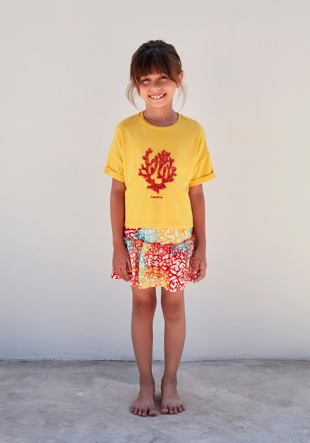 Child standing and smiling in a yellow top with a red coral design, paired with a multicolored coral reef print skirt