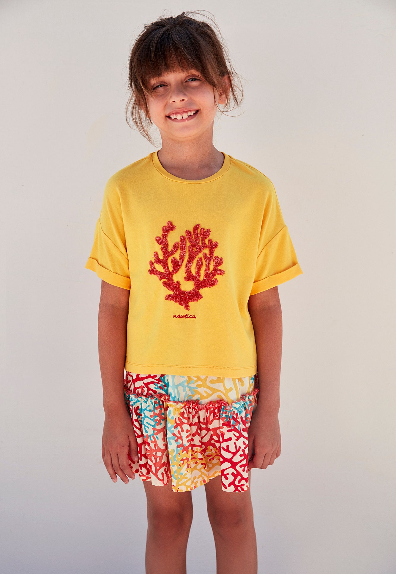 Child wearing a yellow top with a red coral design and a multicolored coral reef print skirt, standing and smiling