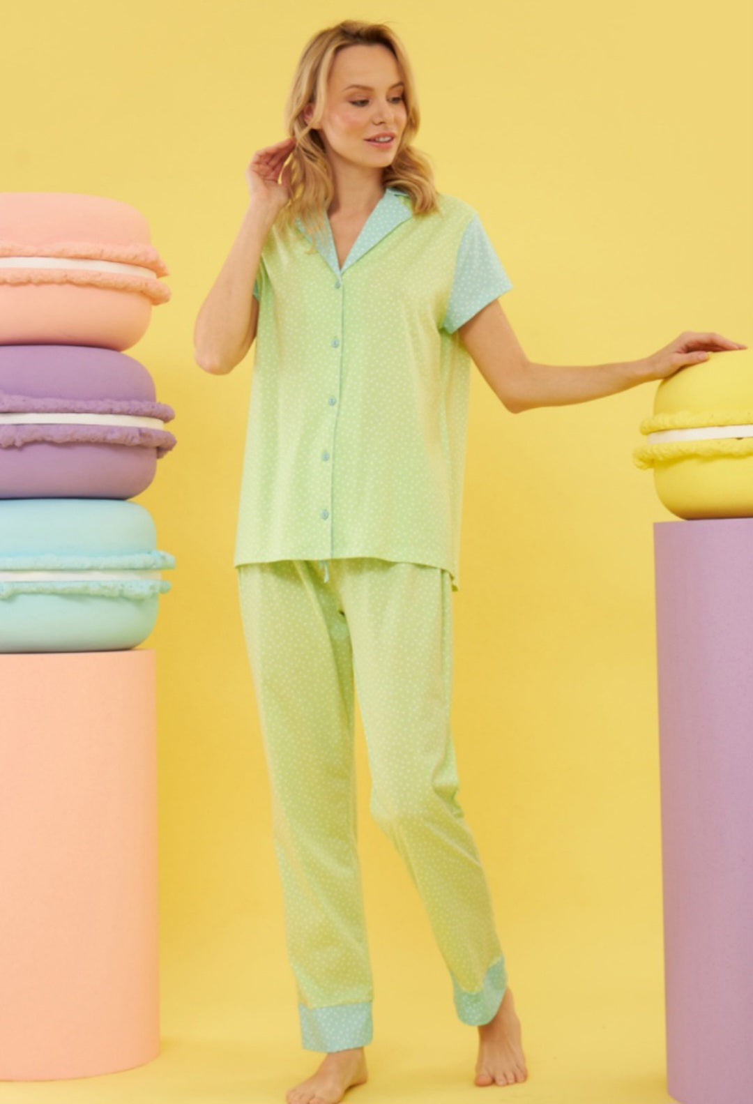 Model wearing the Popjammies "Macaron Dreams" Pajama Set, featuring a soothing green and blue polka dot pattern, standing beside colorful macaron props against a yellow background.