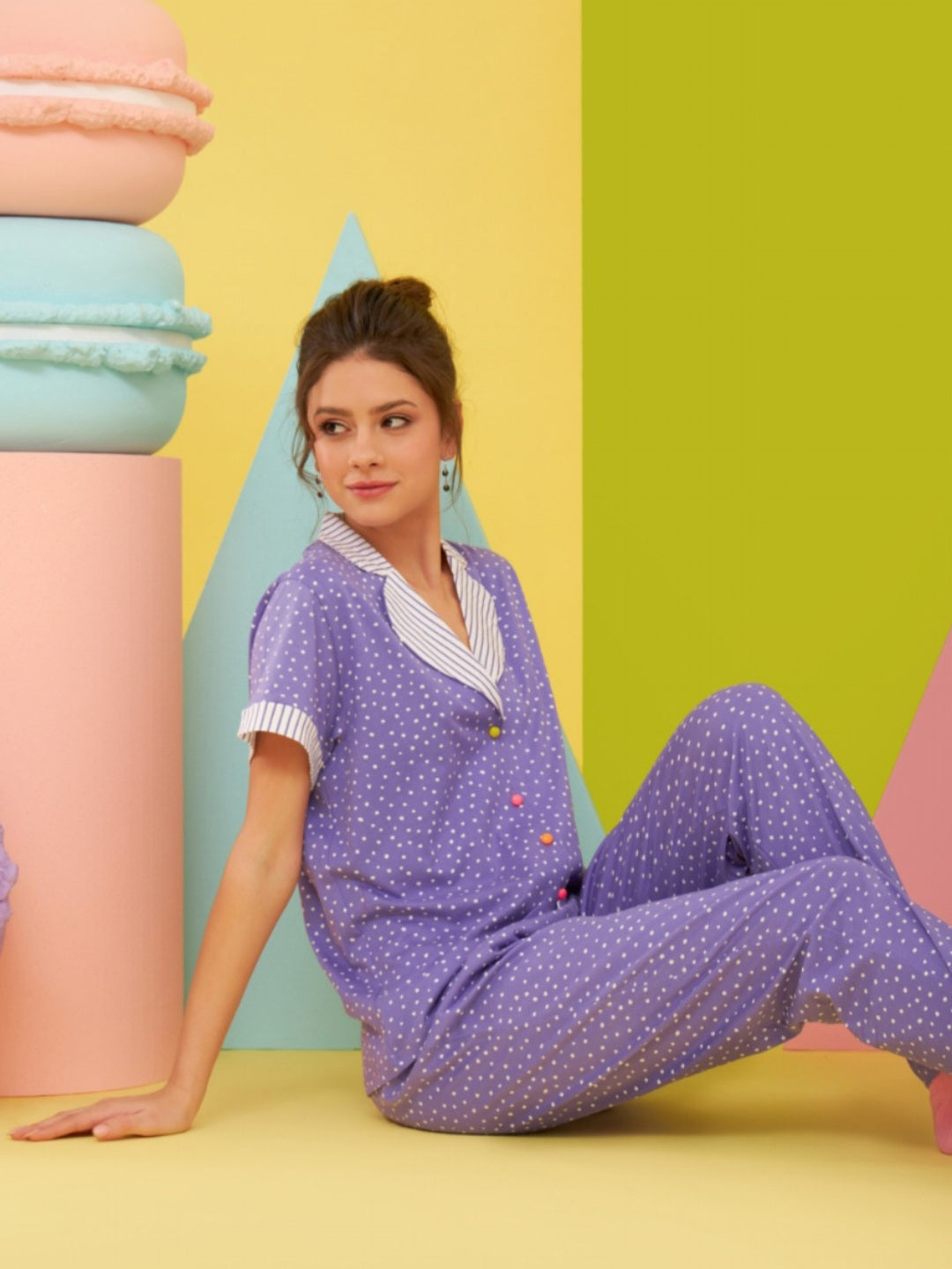 Model wearing the Popjammies "Polka Dot Delight" Pajama Set, featuring a purple top with white polka dots and colorful buttons, and matching pants, sitting against a colorful pastel backdrop.