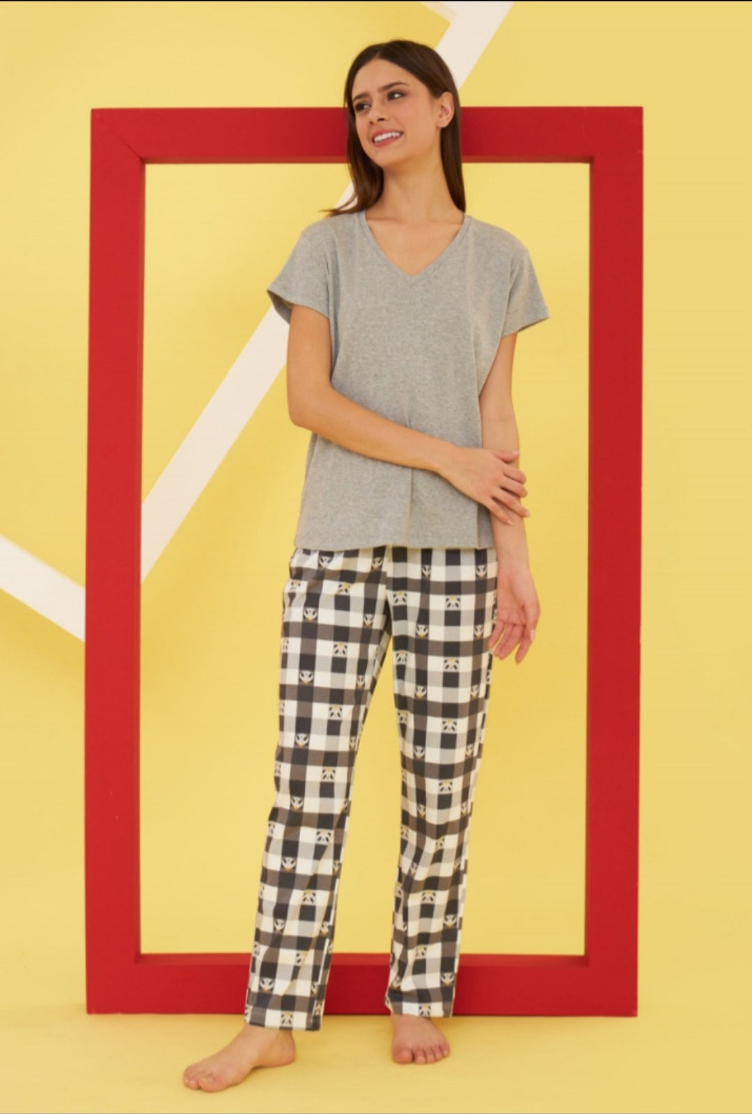 Model wearing the Popjammies "Cozy Check" Pajama Set, featuring a grey V-neck top and black and white checkered pants with panda faces, standing against a bright yellow backdrop with a red frame.