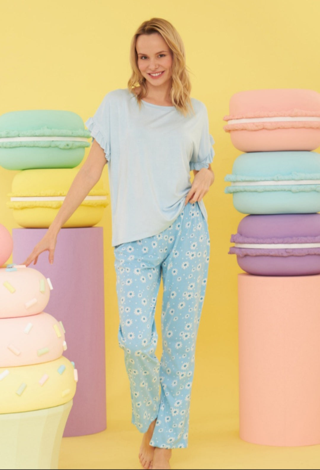 Model wearing the Popjammies "Blue Daisy Dreams" Pajama Set, featuring a light blue top with ruffled sleeves and sky-blue pants with a daisy print, standing against a colorful backdrop with oversized pastel macarons.