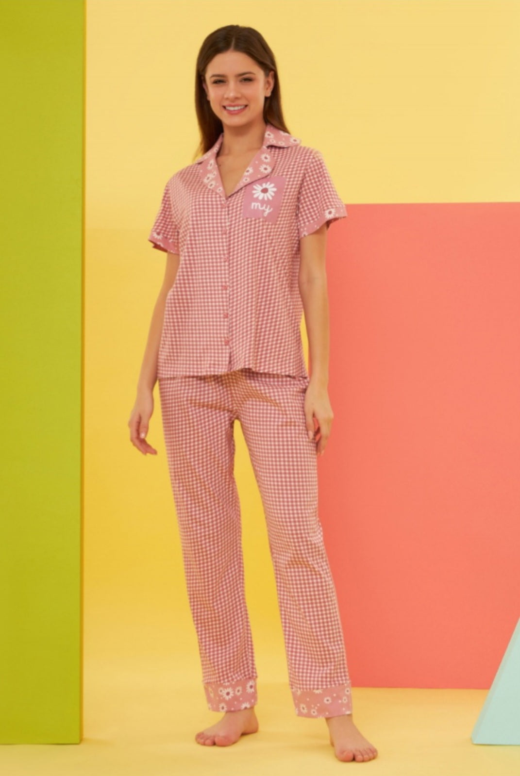 Model wearing the Popjammies "Daisy Delight" Pajama Set, featuring a red and white gingham button-up top with daisy embroidery on the collar and cuffs, and matching pants with daisy-printed cuffs, against a colorful background.