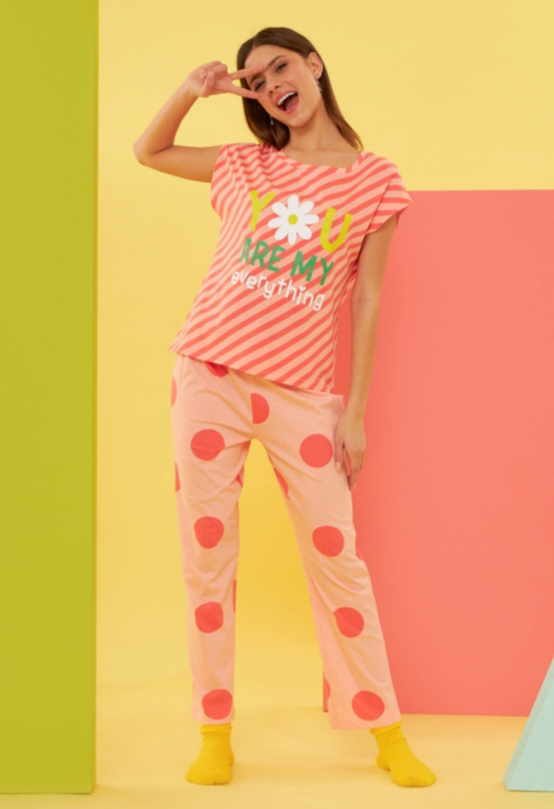 Model wearing the Popjammies "Daisy Delight" Pajama Set, featuring a coral-striped top with "You are my everything" and daisy graphic, paired with pink pants adorned with oversized red polka dots, set against a colorful, playful background.