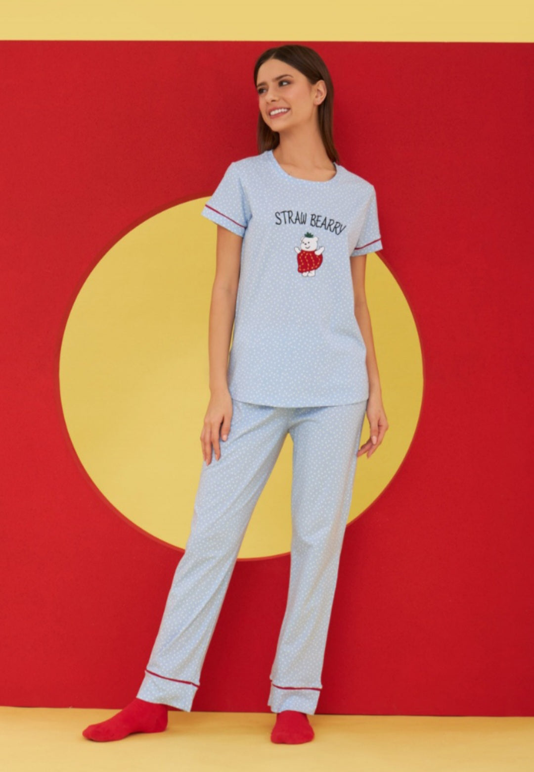 Model wearing the Popjammies "Straw Beary" Pajama Set. The set includes a light blue top with an embroidered bear dressed as a strawberry and matching light blue polka dot pants, set against a vibrant red and yellow background.