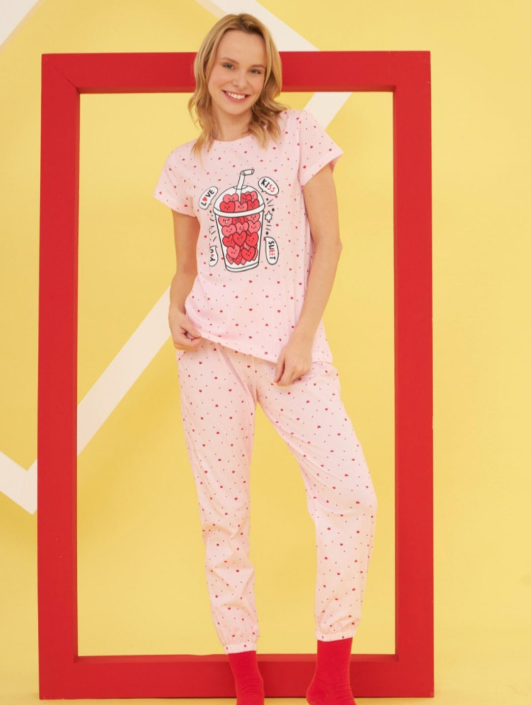 Model wearing the Popjammies "Love Shake" Pajama Set. The set includes a pink top with a love shake graphic and matching pink pants with red heart patterns, set against a playful and colorful background.