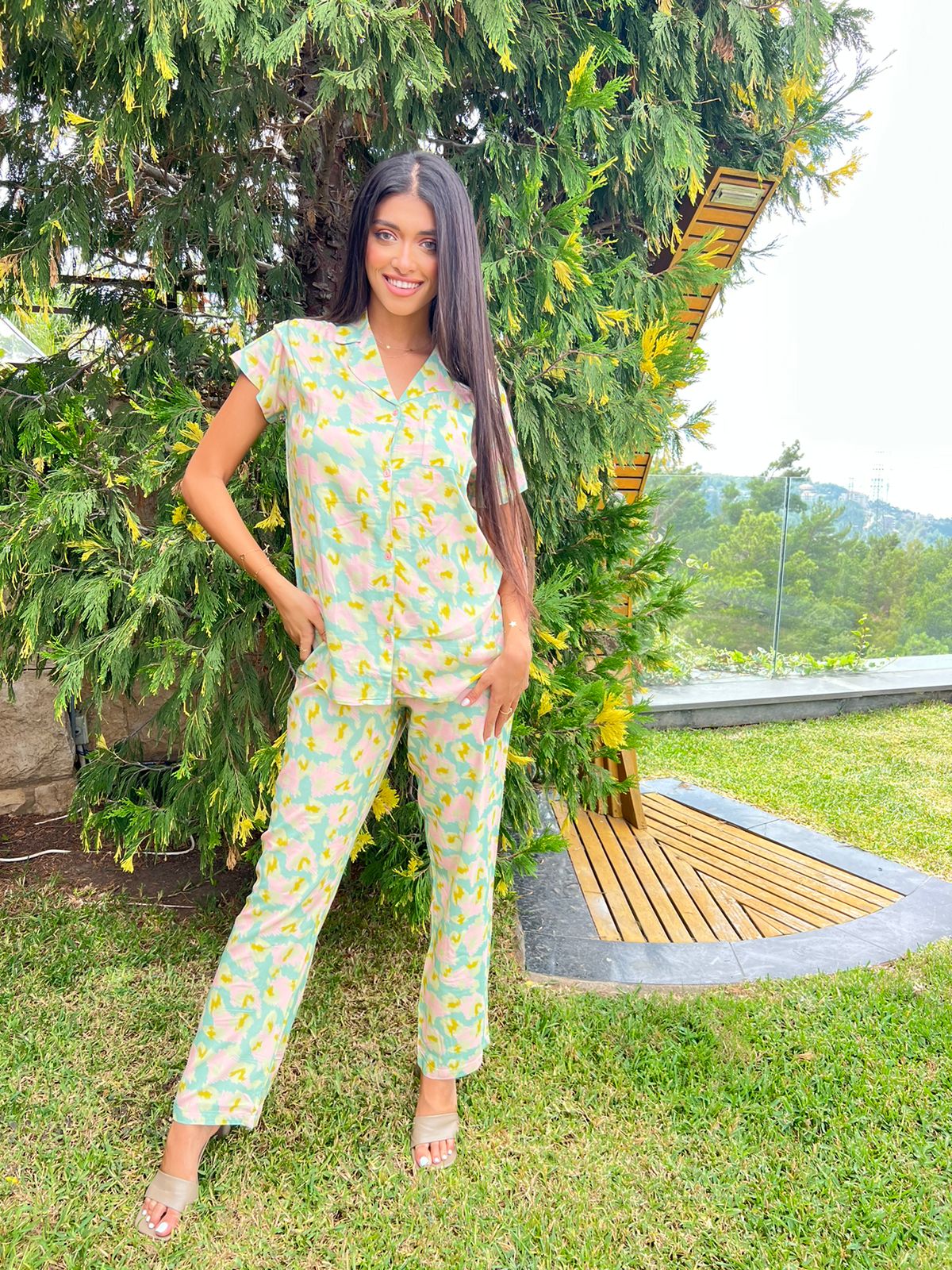 A woman is standing outdoors in front of greenery, wearing a short-sleeved, pastel-colored patterned pyjama set with a notched collar.