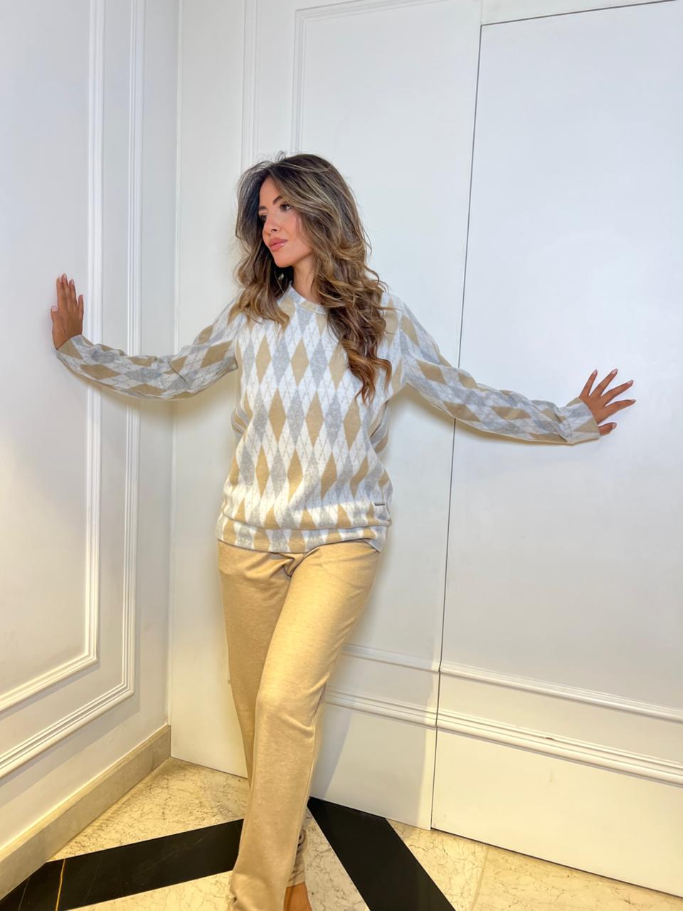 Woman wearing a patterned top and beige pants standing against a white wall.