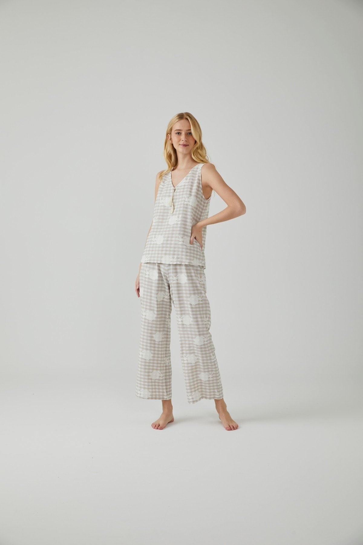 A woman wearing a beige and white gingham patterned pajama set with a sleeveless V-neck top and wide-leg pants, posing elegantly in a minimalist setting.