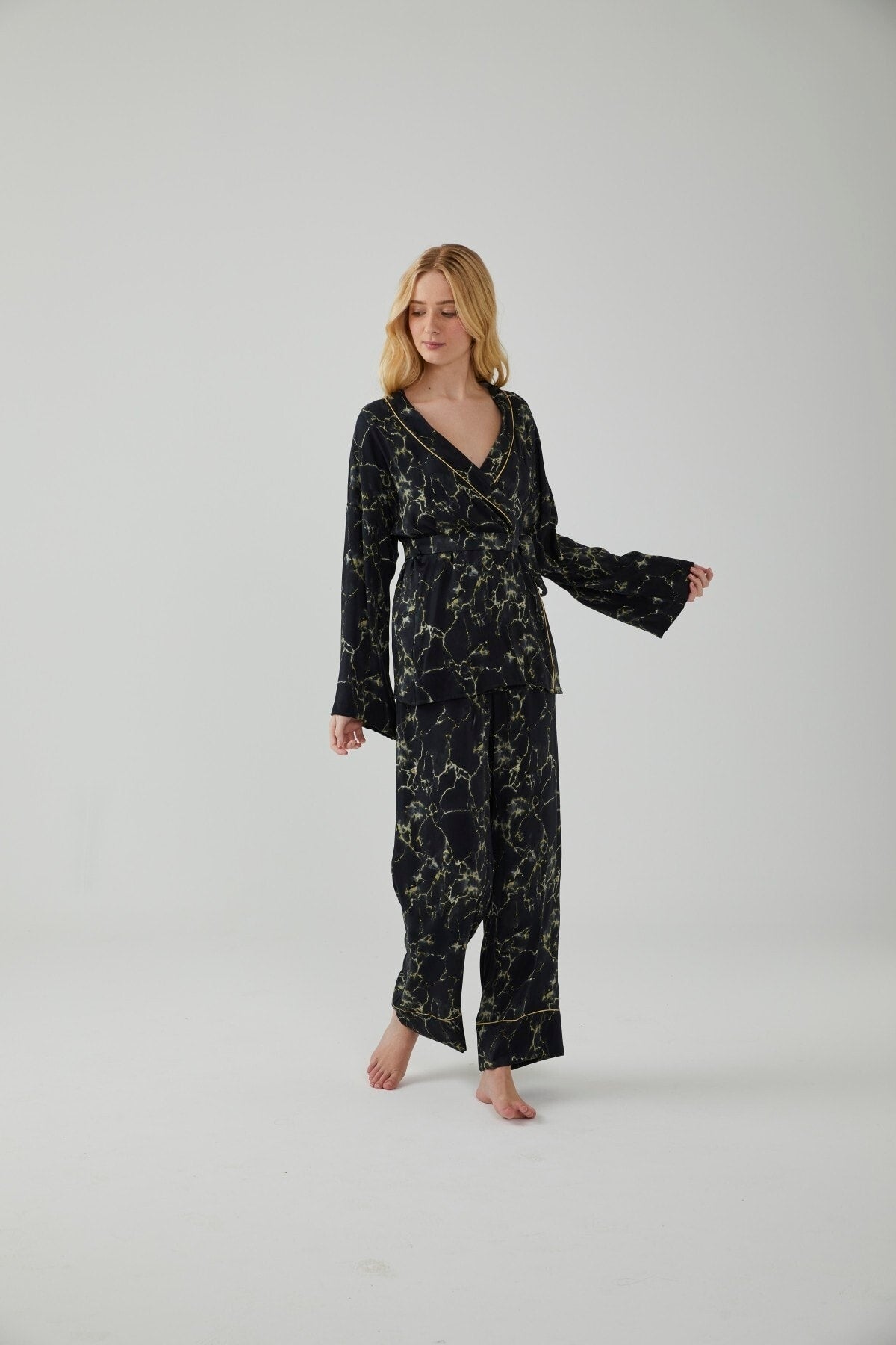 A woman wearing a black and gold marble patterned pajama set with a wrap-style top and wide-leg pants, posing gracefully in a minimalist setting.