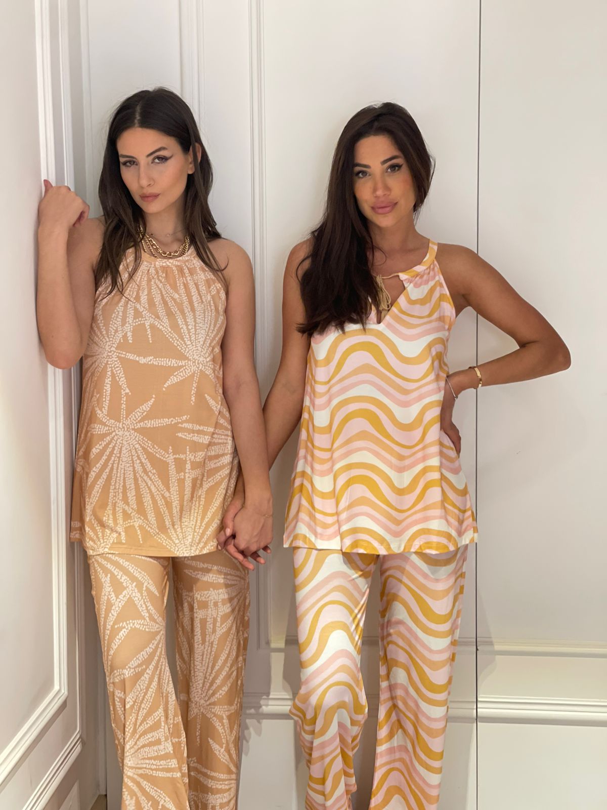 Two women standing side by side in matching pyjama sets from Popjammies. The woman on the left is wearing a beige and white leaf pattern pyjama, while the woman on the right is wearing a pink and yellow wavy stripe pyjama. Both have long hair and are posing against a white wall, holding hands.