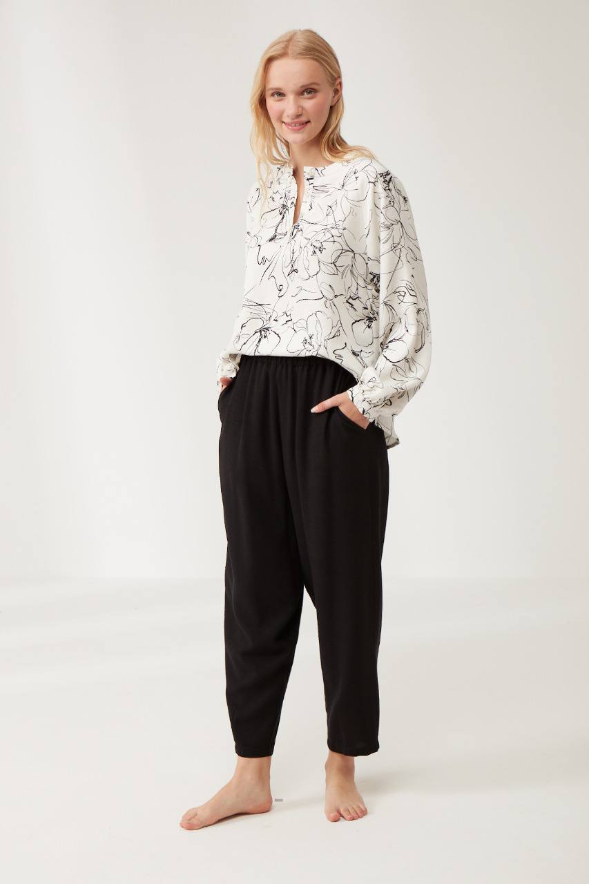 A woman wearing the Popjammies "Chic Comfort" Pyjama Set, featuring a white long-sleeve top with a black floral print and black relaxed pants, standing barefoot in a bright, minimalist setting.