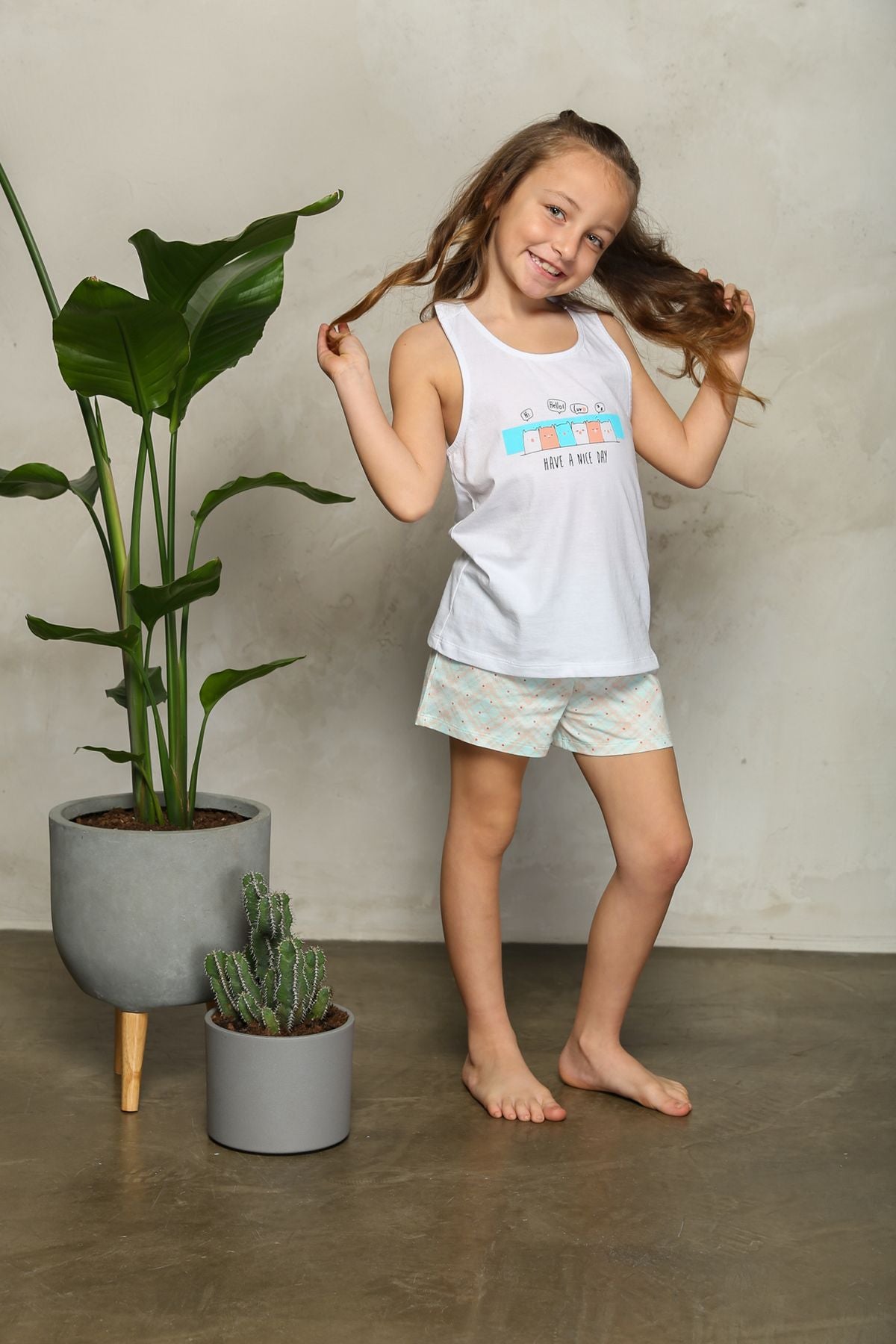 Girl wearing white tank top pajamas with shorts, smiling and playing with her hair.