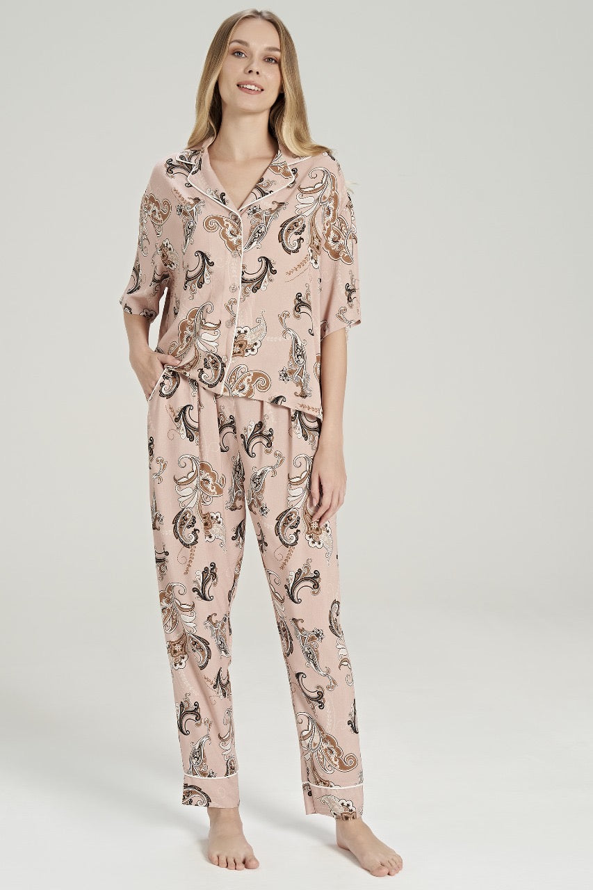 Woman wearing Popjammies Women's Light Pink Paisley Print Pyjama Set. The set includes a short-sleeve, button-down top with a notched collar and matching full-length pants. Both pieces feature an intricate paisley pattern in shades of brown, cream, and black on a light pink background.