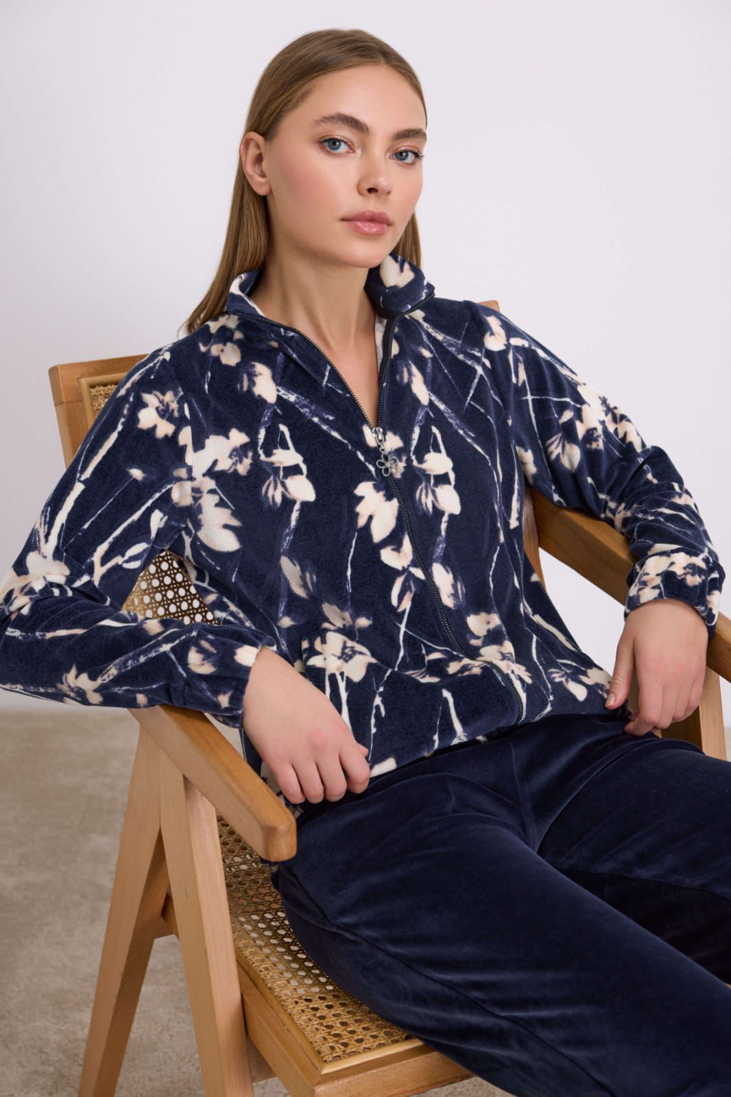 Woman wearing a navy floral blouse and dark pants sitting on a wooden chair.