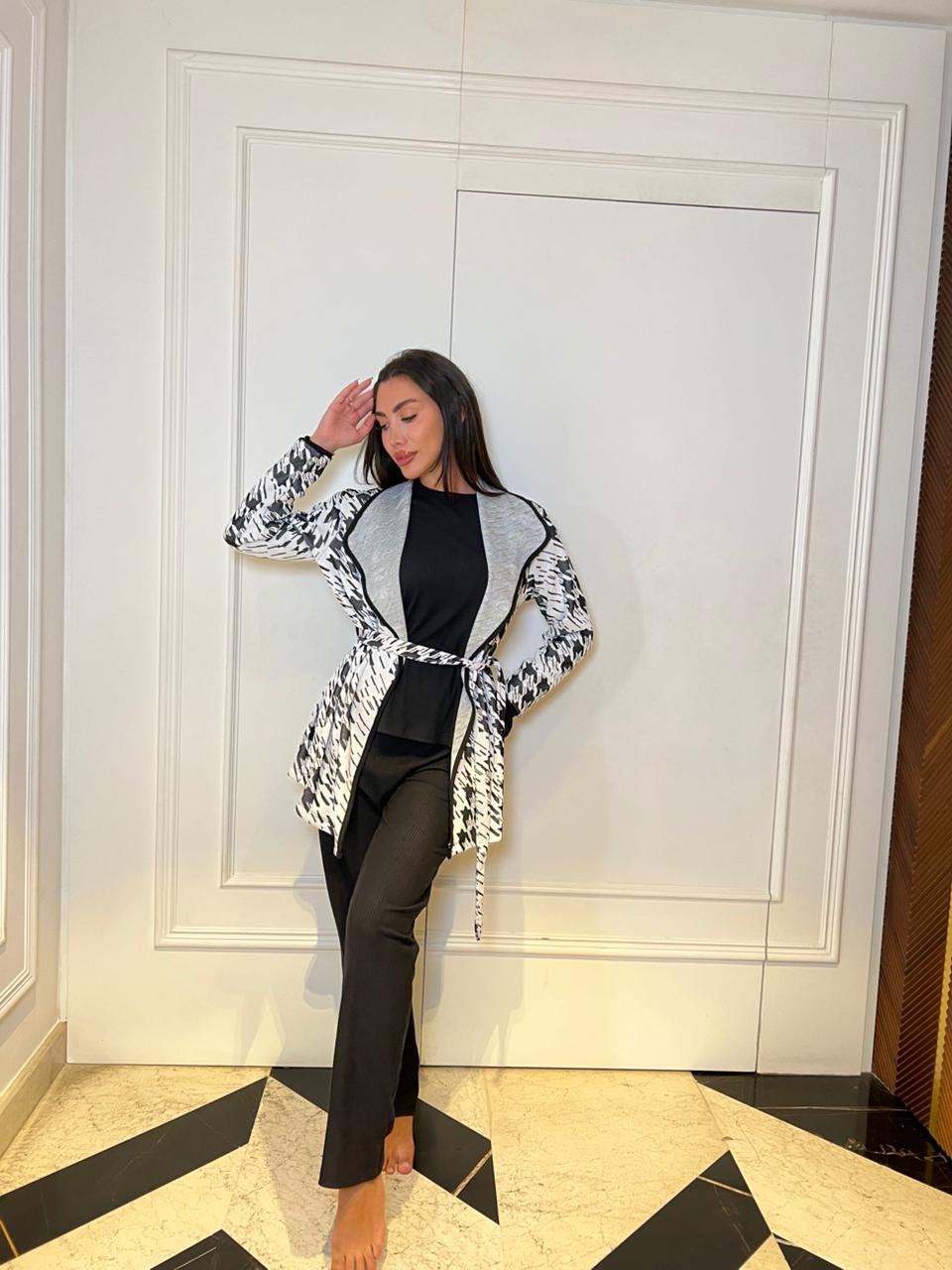 Woman posing in a stylish outfit with a patterned cardigan and black pants against a white wall.