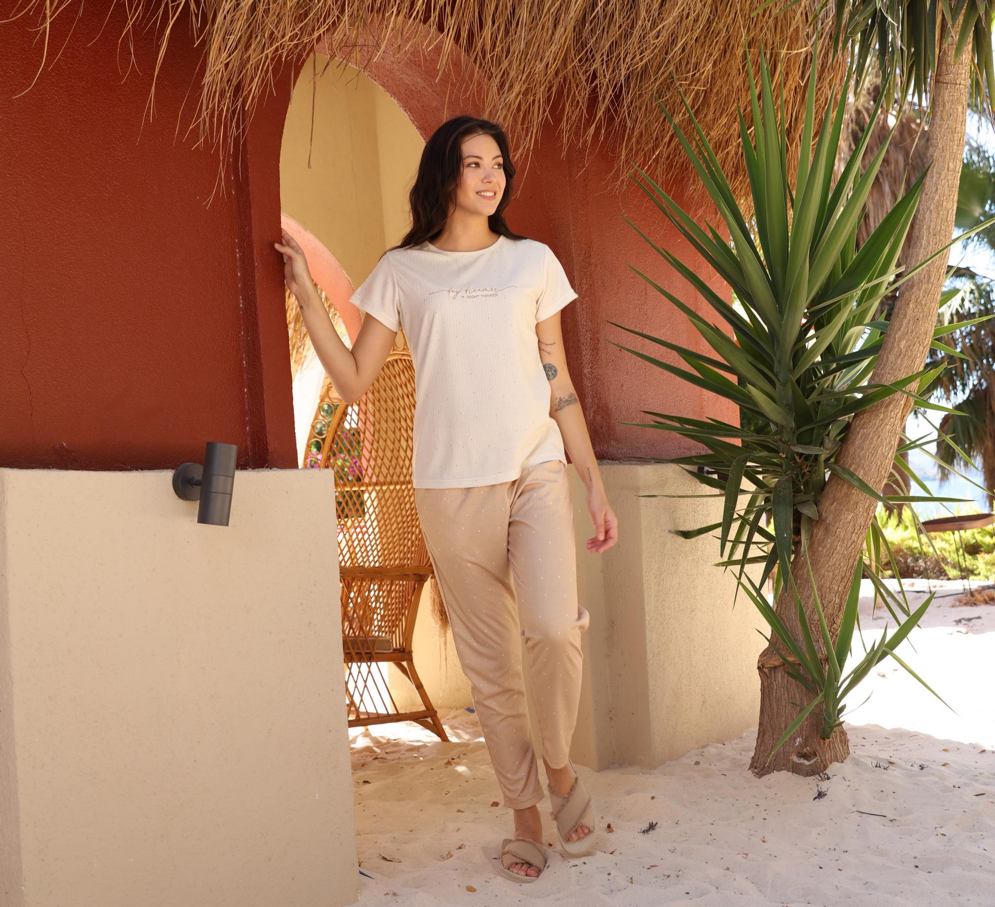 Woman wearing Popjammies "Desert Oasis" Pyjama Set, featuring a creamy top and beige pants with shimmering details, standing in a cozy, tropical outdoor setting.