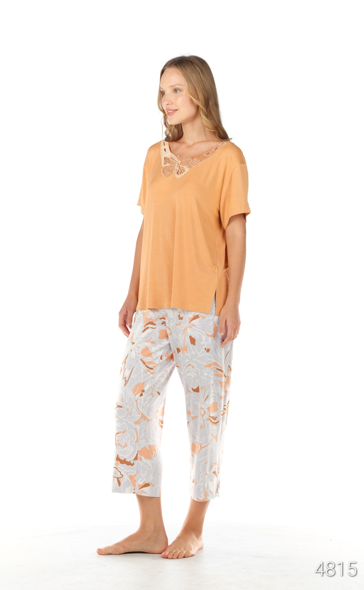 Woman standing and wearing a peach short-sleeved top with a butterfly lace neckline and white floral patterned pants from the Popjammies Women's Butterfly Lace Pyjama Set.