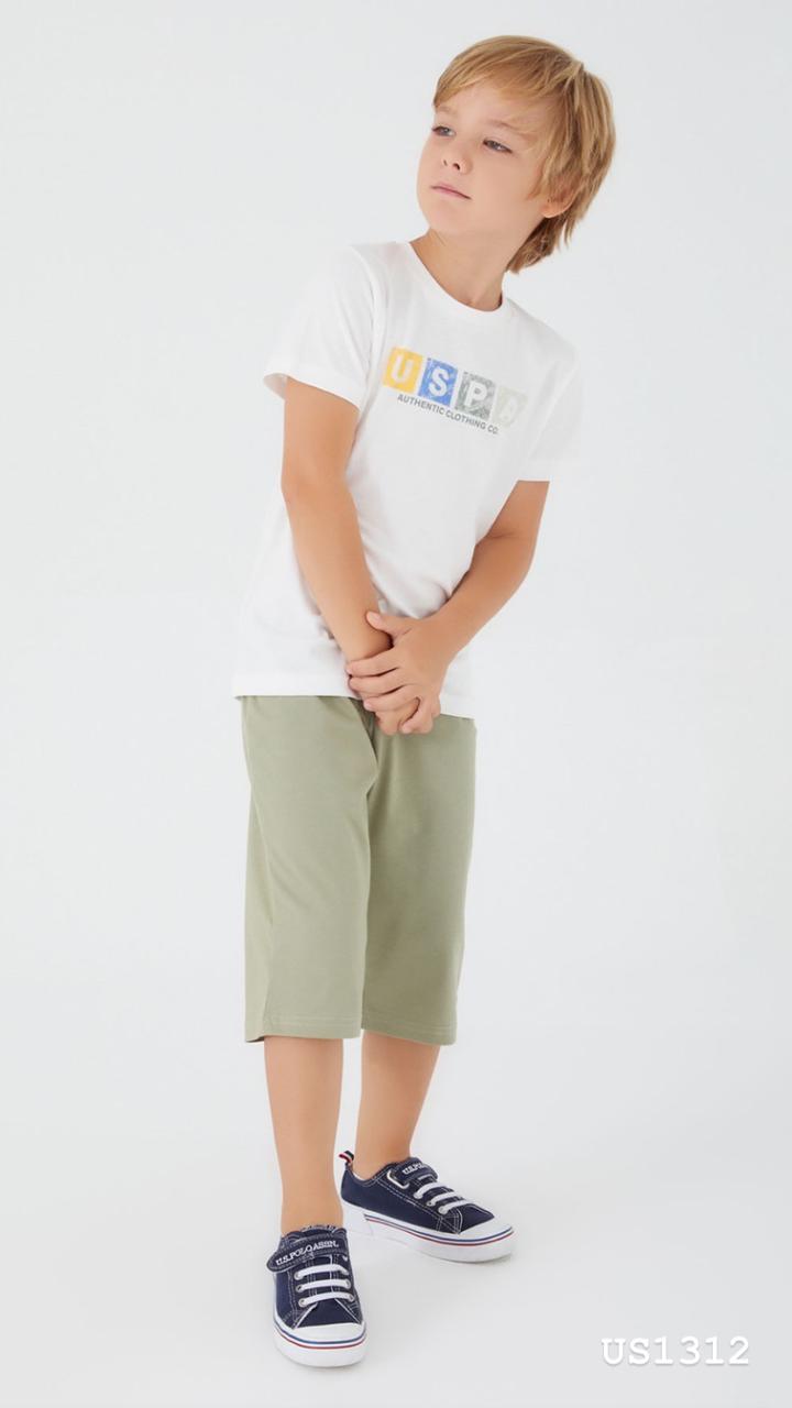 Boy wearing Popjammies pyjama set with a white tee featuring a colorful "USPA Authentic Clothing Co." logo and olive green shorts, standing with hands clasped.