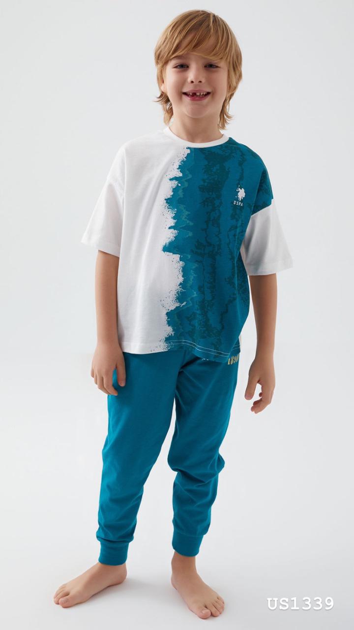 Boy wearing a white top with a blue graphic and blue pants, standing barefoot and smiling.