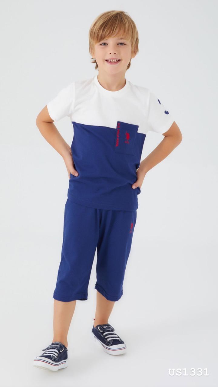 Boy wearing a white and navy color block top with a front pocket and navy pants, standing and smiling.