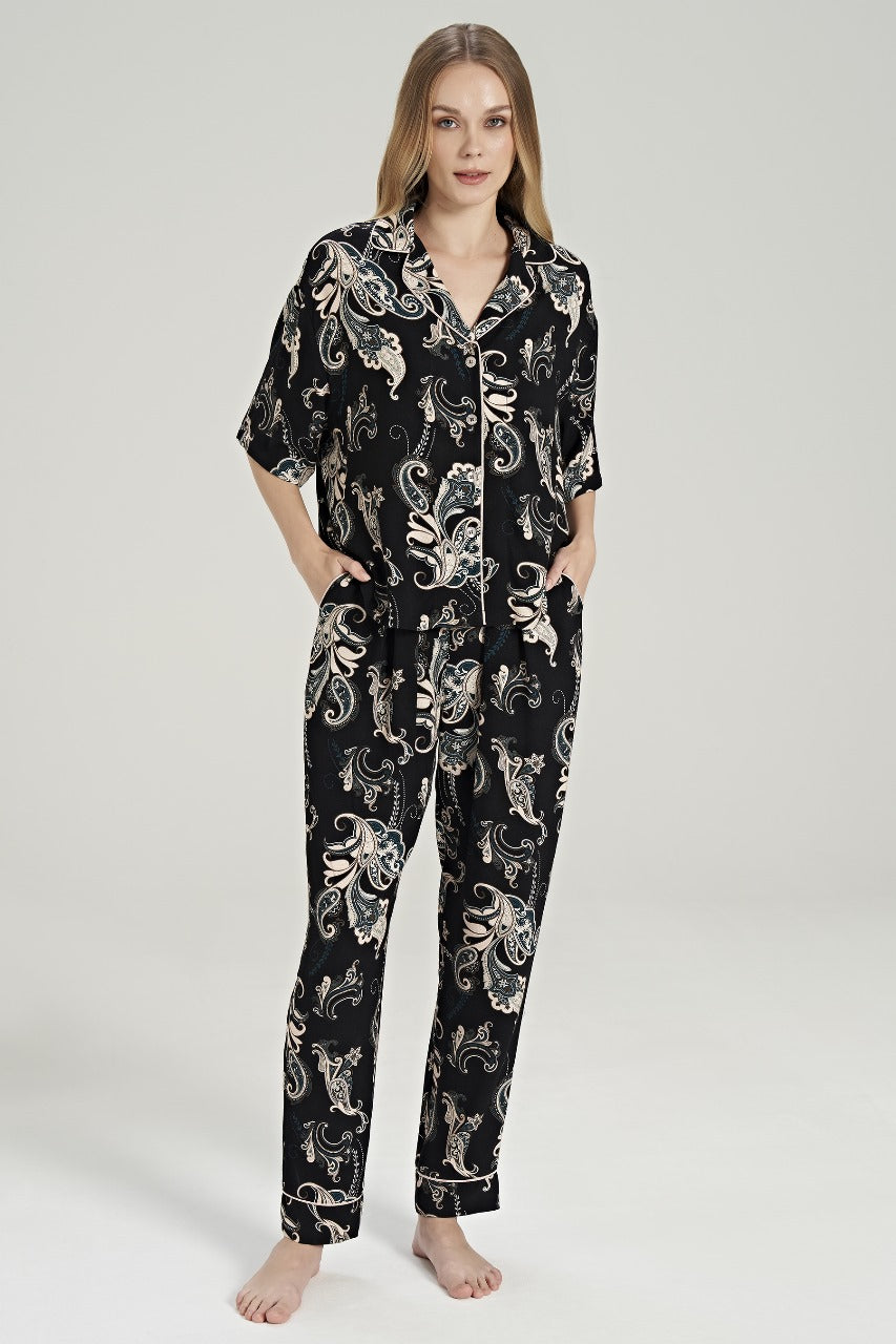 Woman wearing Popjammies Women's Black Paisley Print Pyjama Set. The set includes a short-sleeve, button-down top with a notched collar and matching full-length pants. Both pieces feature an intricate paisley pattern in shades of cream, beige, and dark teal on a black background.