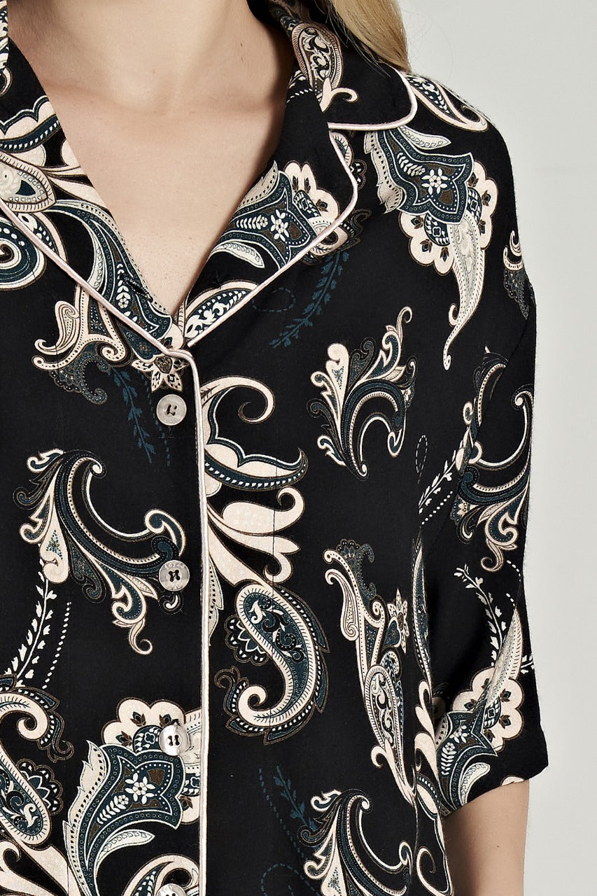Close-up of the Popjammies Women's Black Paisley Print Pyjama Top, showcasing intricate paisley patterns in cream and teal on a black background. The top features a classic notched collar with white piping and a button-down front.