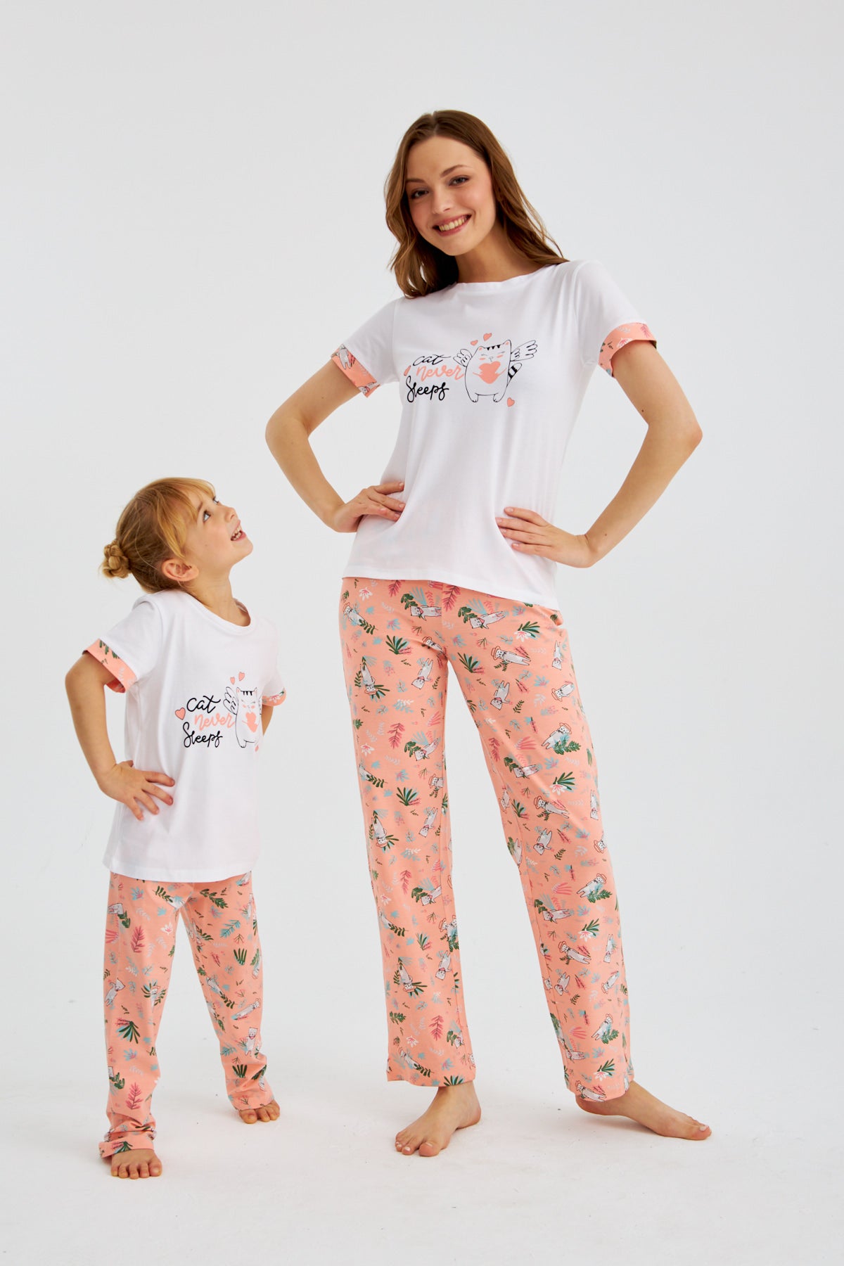  Mother and daughter posing in matching Popjammies "Cat Never Sleeps" pyjama set, hands on hips, showcasing the white tops with cat graphic and pink floral print pants.