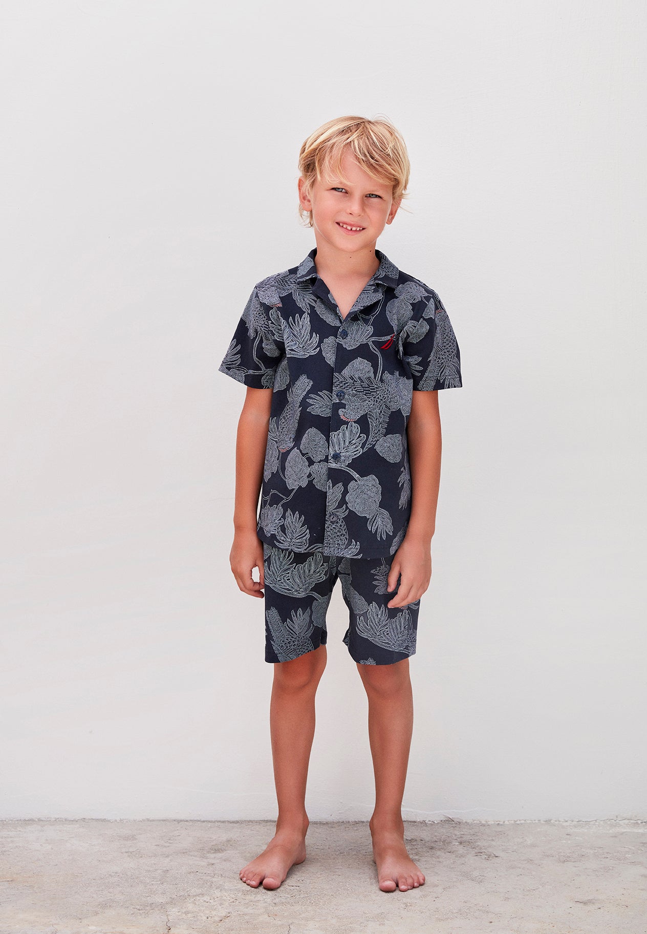 Full body view of a boy in a navy blue nautical pajama set with ocean-inspired patterns, standing barefoot.