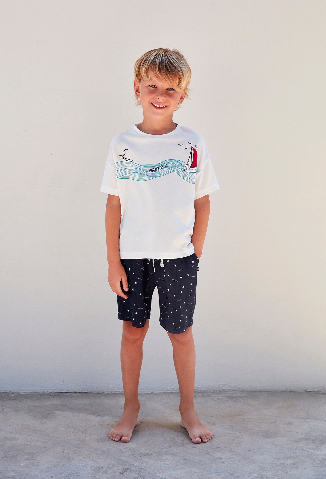 Boy wearing a white top with a boat and wave print and navy blue shorts from Popjammies, standing against a white background.