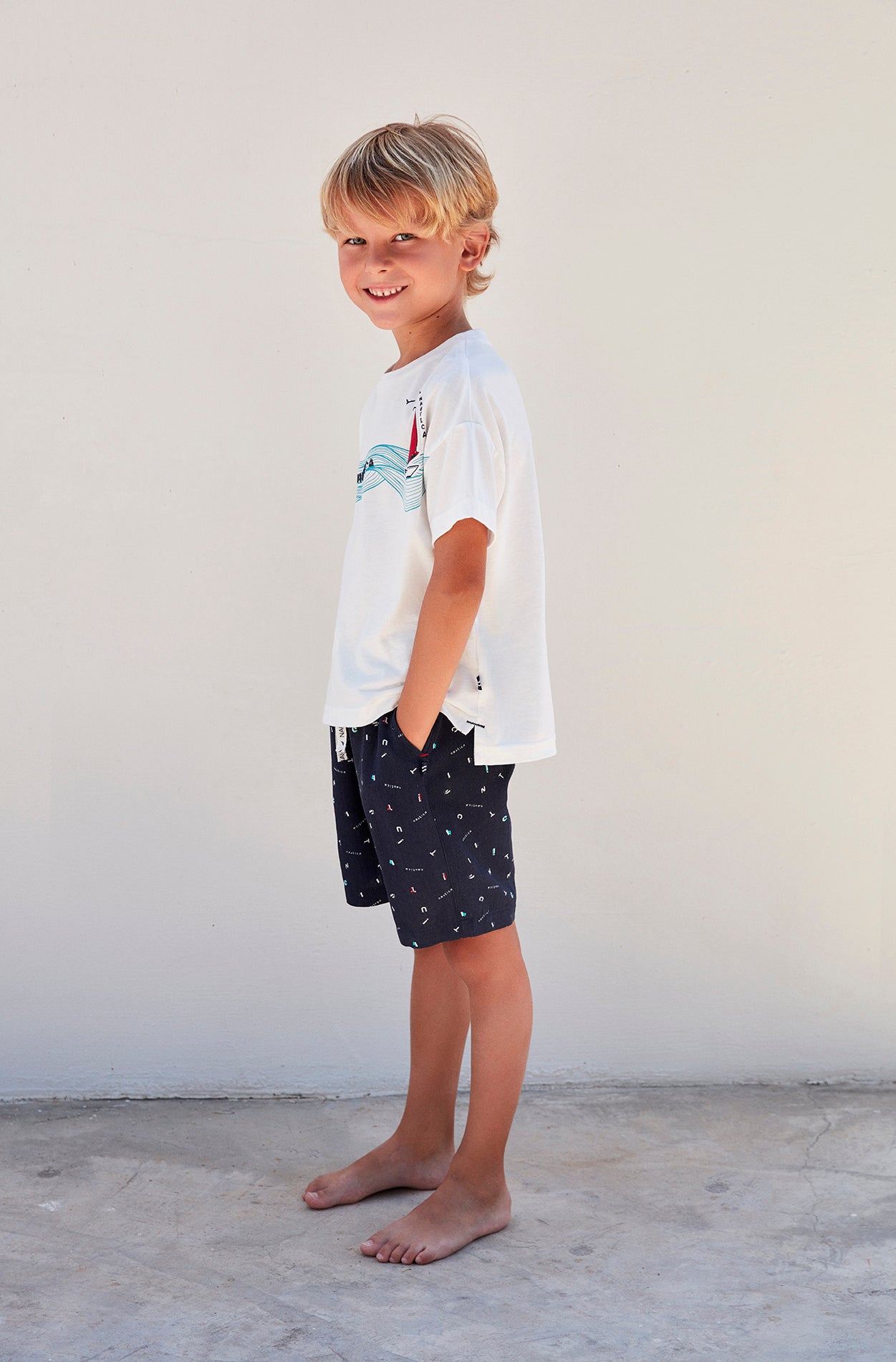 Side view of a boy wearing a white top with a boat and wave print and navy blue shorts from Popjammies, standing against a white background