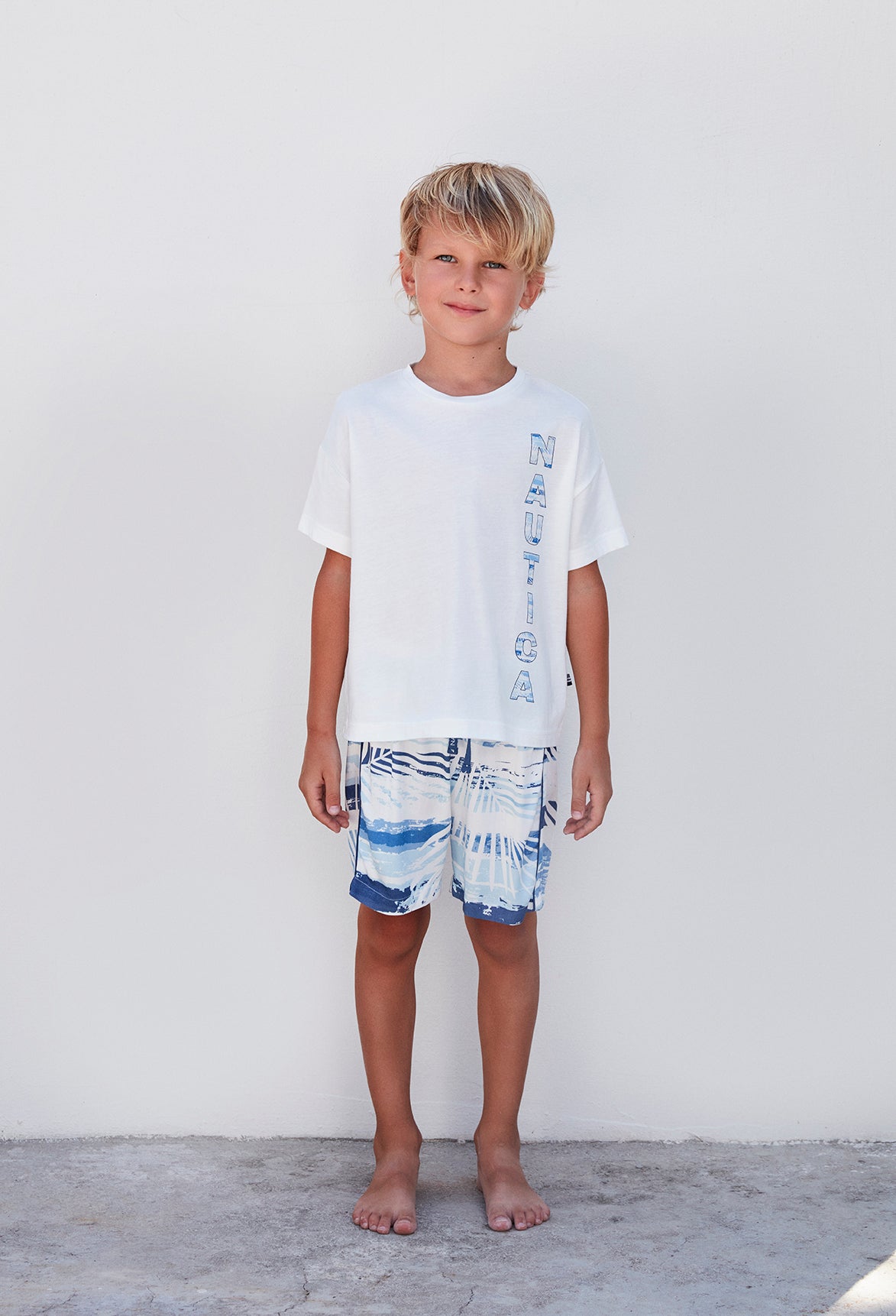 Full-body view of a boy in Popjammies Nautica pajama set featuring a white t-shirt and wave-patterned shorts.