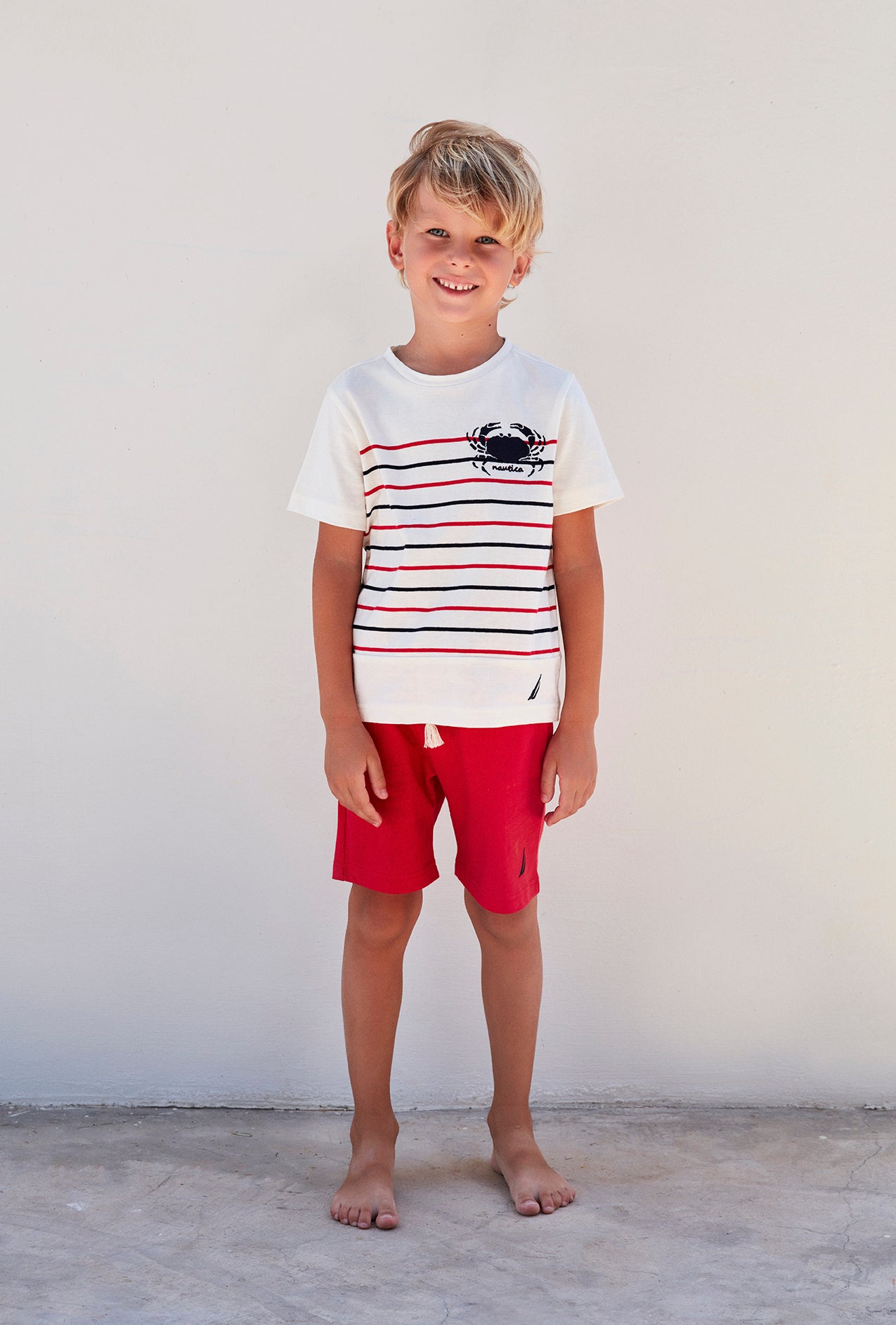 Boy smiling in white top with red and navy patterns and crab emblem, and red shorts from Popjammies