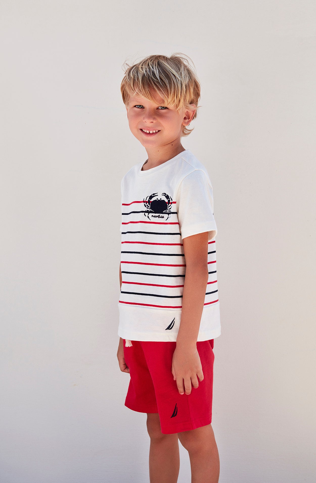 Side View of a boy smiling in white top with red and navy patterns and crab emblem, and red shorts from Popjammies
