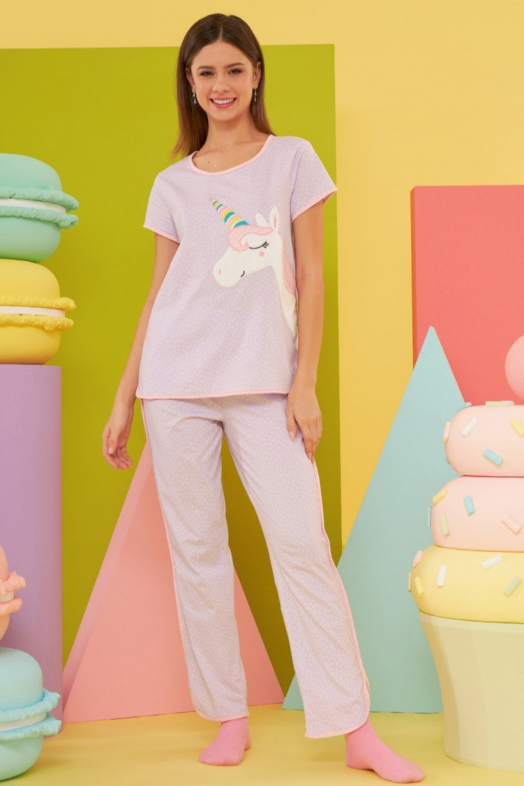 Model wearing the Popjammies "Unicorn Dream" Pajama Set, featuring a lilac top with a whimsical unicorn graphic and matching lilac pants with white polka dots, standing against a colorful pastel backdrop.