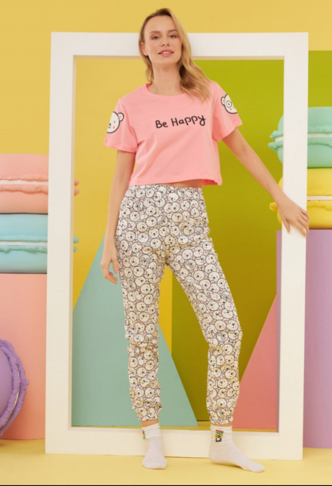 Model wearing the Popjammies "Be Happy" Pajama Set. The set includes a pink crop top with "Be Happy" written on it and pants adorned with a cheerful smiley face print, set against a colorful, playful background.
