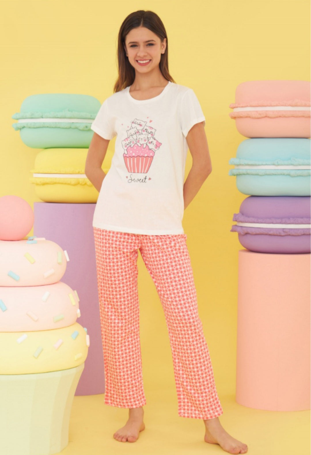 Model wearing the Popjammies "Sugar Dreams" Pajama Set. The set includes a white top with a cupcake graphic and the word "Sweet," paired with red gingham pants. The bright, pastel-colored background adds to the cheerful and playful nature of the pajamas.