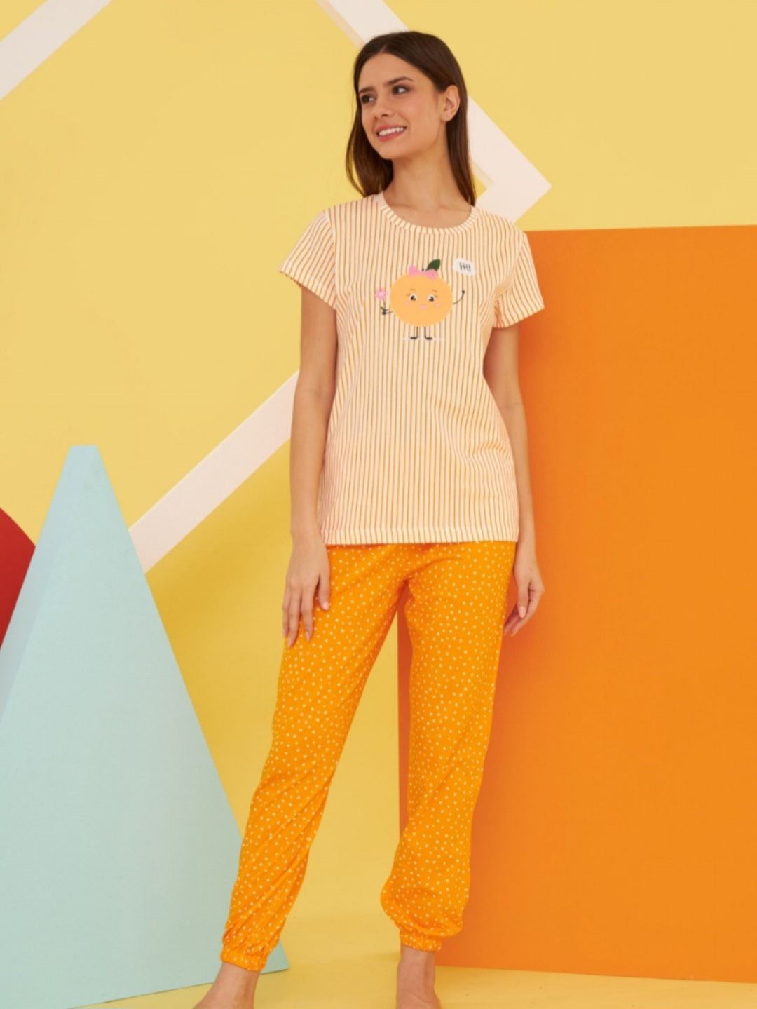 Model wearing the Popjammies "Sunny Delight" Pajama Set. The set includes a striped top with a cartoon orange character and matching orange polka dot pants. The bright background enhances the cheerful and playful design of the pajamas.