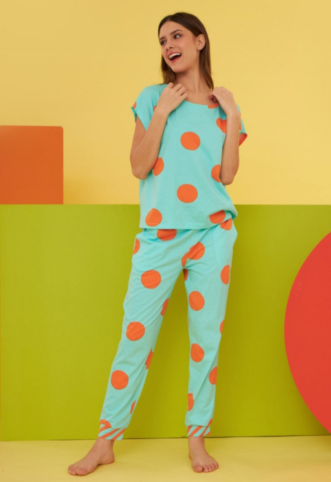Model wearing the Popjammies "Playful Polka Dot" Pajama Set. The set is aqua with large orange polka dots, including a loose-fitting top and matching pants with striped cuffs. The bright and colorful background complements the vibrant design, highlighting the fun and cheerful nature of the pajamas.