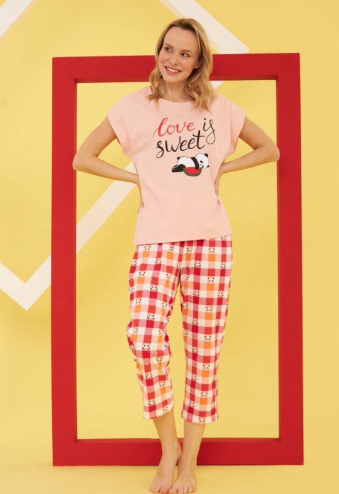 Model wearing the Popjammies "Love is Sweet" Pajama Set. The set includes a pastel pink top with a panda graphic and the words "Love is Sweet," paired with checkered pants in red, pink, and orange featuring panda faces. The bright yellow and red background adds a cheerful and playful atmosphere, highlighting the fun and cozy nature of the pajamas.