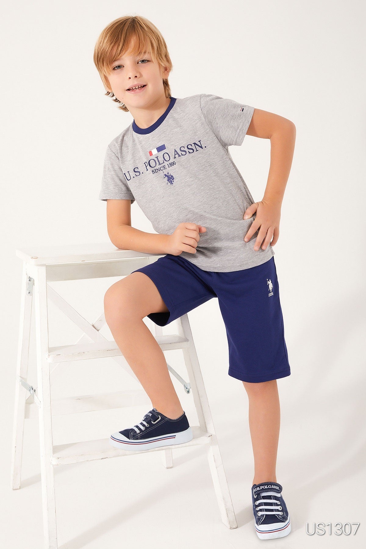 Boy posing beside white chair wearing a grey top with "US Polo Assn" written on it and navy blue shorts from Popjammies.