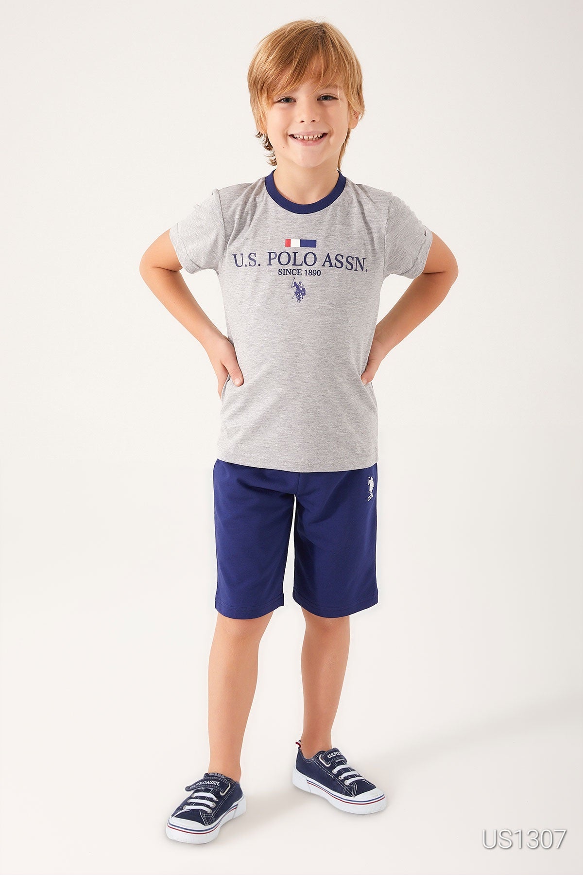  Boy in grey top with "US Polo Assn" print and navy blue shorts, standing with hands on hips.