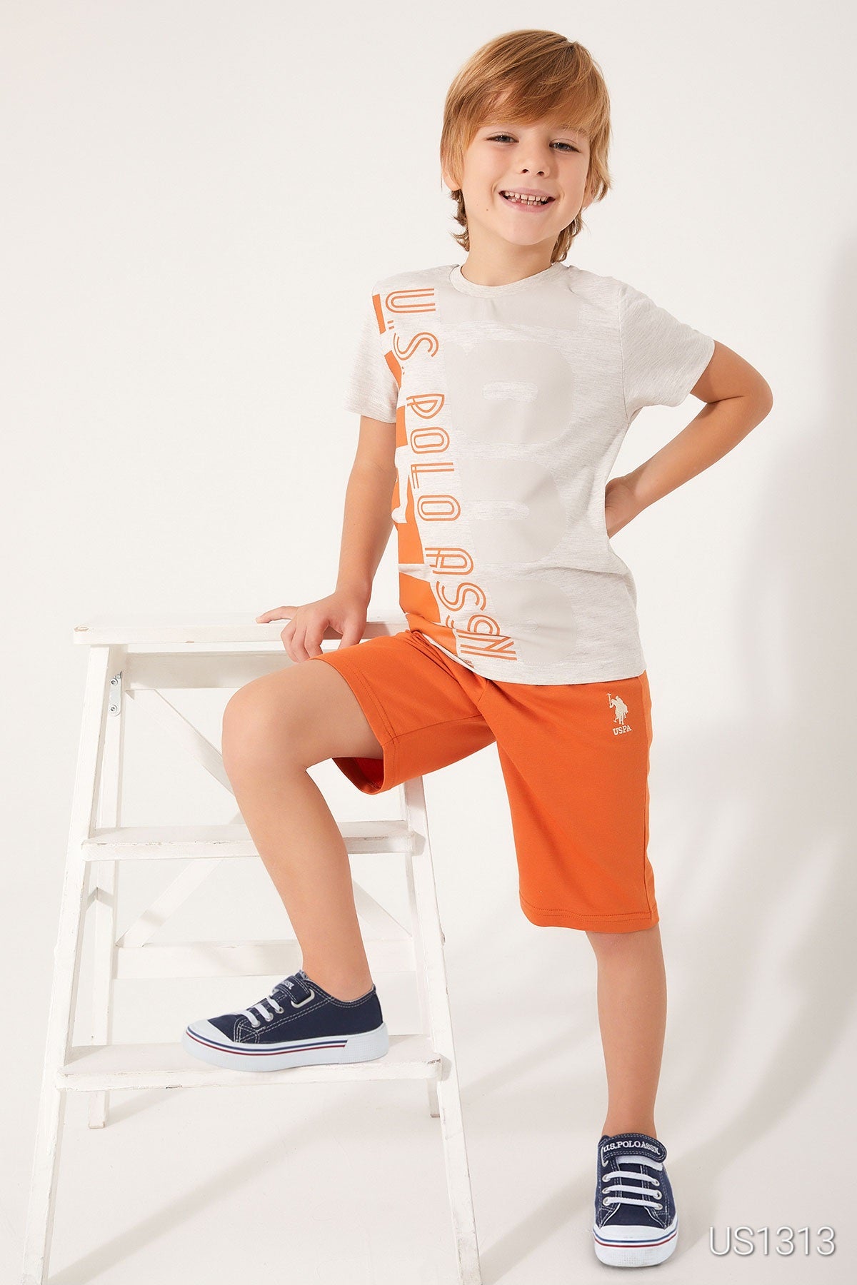 Boy wearing Popjammies pyjama set with a white tee featuring "US Polo Assn." in orange and orange shorts, standing beside a white chair.