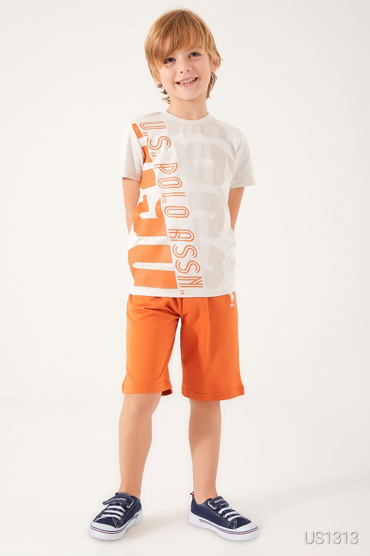 Boy wearing Popjammies pyjama set with a white tee featuring "US Polo Assn." in orange and orange shorts, standing with hands behind his back.