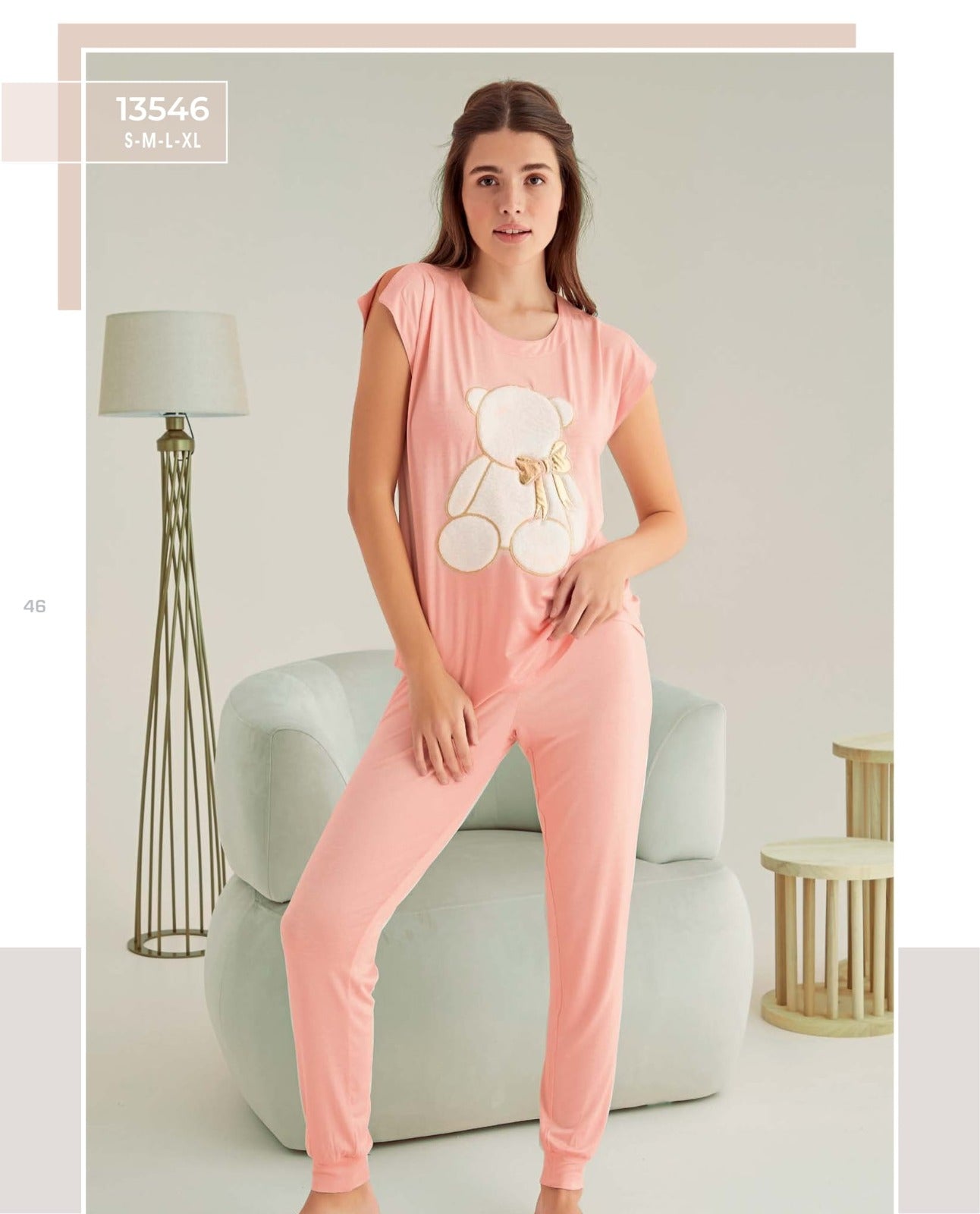 Woman wearing a pink pyjama set with a teddy bear applique, featuring short sleeves and elasticized cuffs, standing in a cozy room.