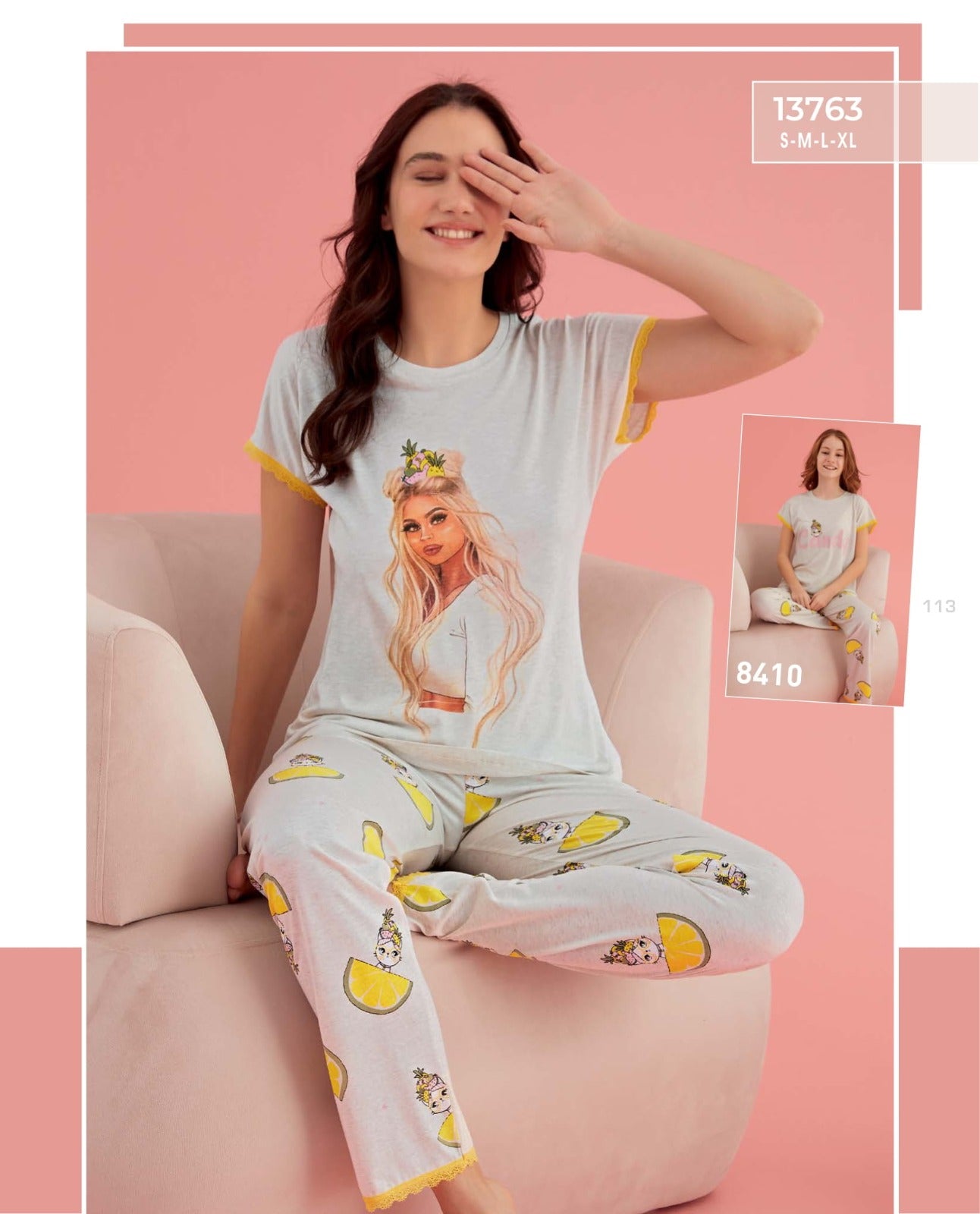 A woman sitting on a pink sofa, wearing a grey pyjama set with a graphic print of a stylish woman on the top and fun lemon and pineapple prints on the pants.
